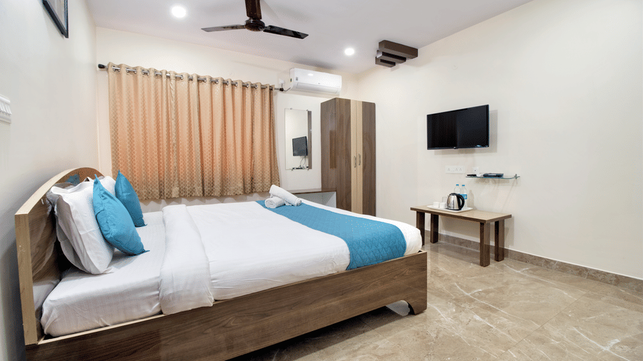 Spacious hotel room featuring a neatly made bed with blue runners, a wall-mounted television, and a simple wooden desk area at beestay Hotels, MVP Colony, Vizag.