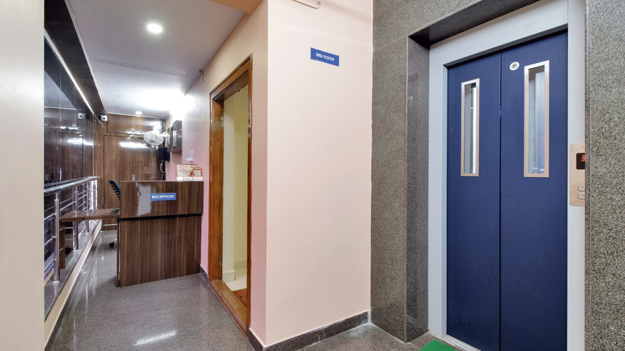 A modern hotel corridor featuring a lift with blue doors, a small wooden reception desk, and bright, recessed ceiling lights at beestay Hotels, MVP Colony, Vizag.