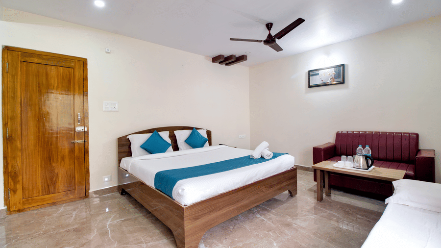 A spacious double bed with blue runners and pillows in a bright room featuring minimalist wooden furniture and a ceiling fan at beestay Hotels, MVP Colony, Vizag.