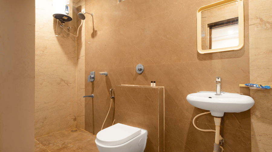 A clean, compact hotel bathroom with beige tiled walls, a white ceramic toilet, a pedestal sink, and a wall-mounted mirror at beestay Hotels, MVP Colony, Vizag.