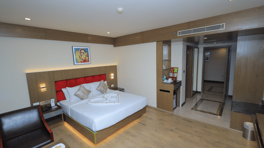 Deluxe Room at Benzz Park, Vellore. Queen bed with red headboard, towel heart, wood floor, lounge chair, and a view of the adjacent entryway.