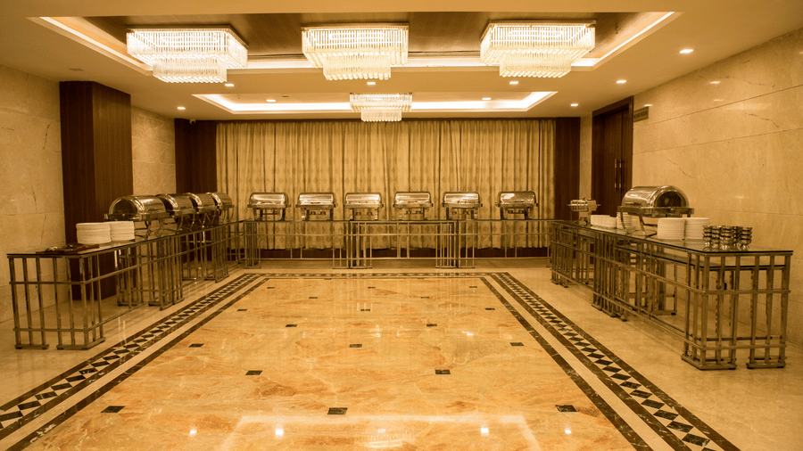 A brightly lit banquet hall with several chafing dishes lined at the far end of the hall at Benzz Park, Vellore.