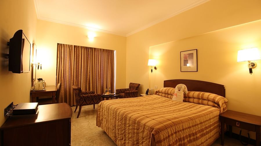 A room with a large bedding with wall lamps on either side of the bed, coffee table and chairs, on a bright lighted setting.