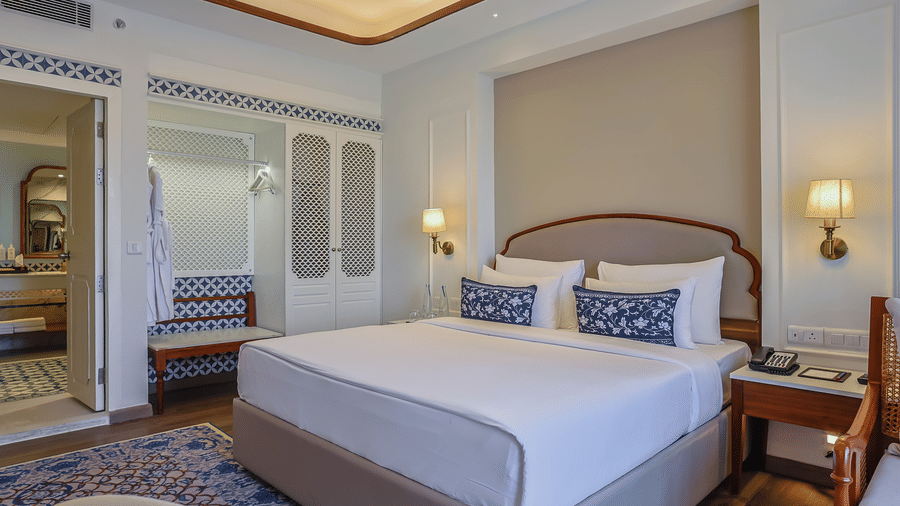 Spacious hotel bedroom featuring a king bed with blue accent pillows, built-in wardrobe storage, warm cove lighting, and an adjoining bathroom at Bhagirathi by Atishay.
