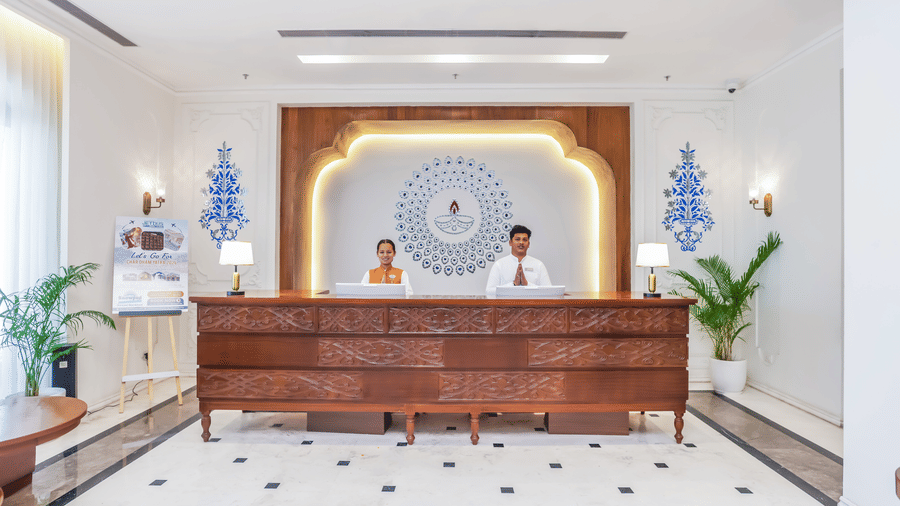 Two reception staff members standing behind a long wooden front desk with an illuminated arched wall backdrop at Bhagirathi by Atishya.