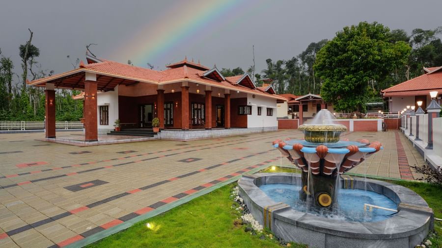 The traditional facade of Bhuthanakadu Retreat under a vibrant rainbow, featuring a fountain in a garden and a colorful patterned walkway leading to the main buildings.
