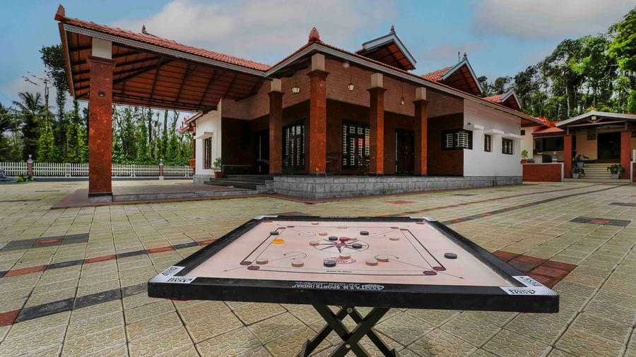 A carrom board is set up outdoors on a paved courtyard, with the traditional architecture of Bhuthanakadu Retreat visible in the background.