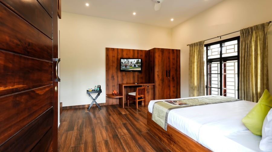 A wide-angle view of the spacious premium room at Bhuthanakadu Retreat, showcasing a large bed, a single bed, a TV on the wall, and rich wooden flooring.
