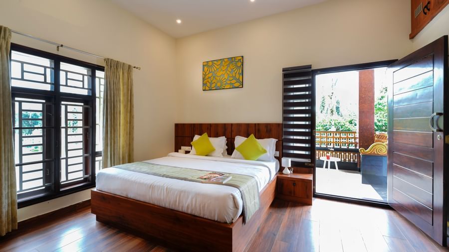  A premium room at Bhuthanakadu Retreat, featuring a large wooden bed with bright green and white bedding, and a sliding glass door leading to a balcony.