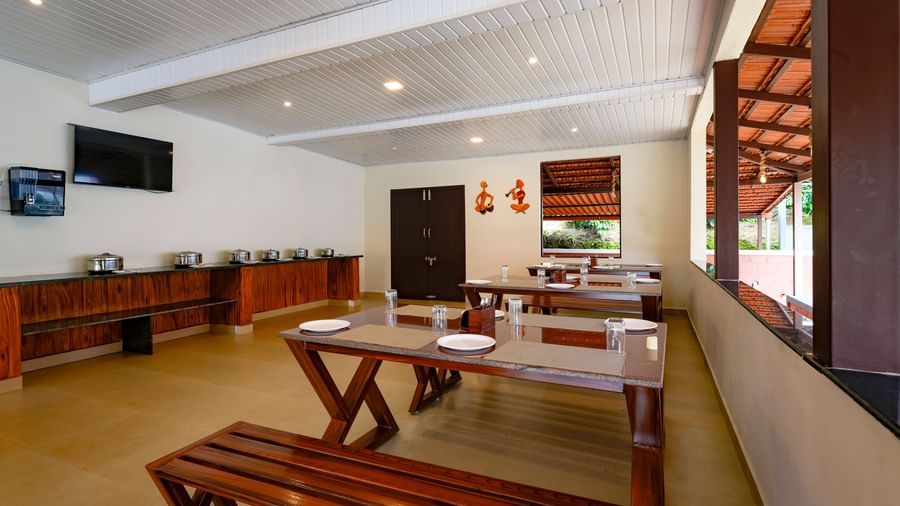 A daytime interior view of the Mane Cafe at Bhuthanakadu Retreat, highlighting a buffet area with covered dishes, a TV on the wall, and long wooden dining tables with benches.