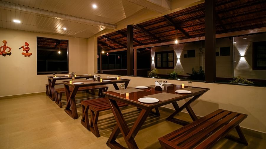 An evening shot of the Mane Cafe at Bhuthanakadu Retreat, showing a well-lit, open-air dining space with rustic wooden tables and bench seating.
