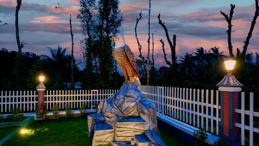 A wide shot of the illuminated eagle statue and manicured garden at Bhuthanakadu Retreat, under a dramatic, colorful evening sky with shades of pink and orange.