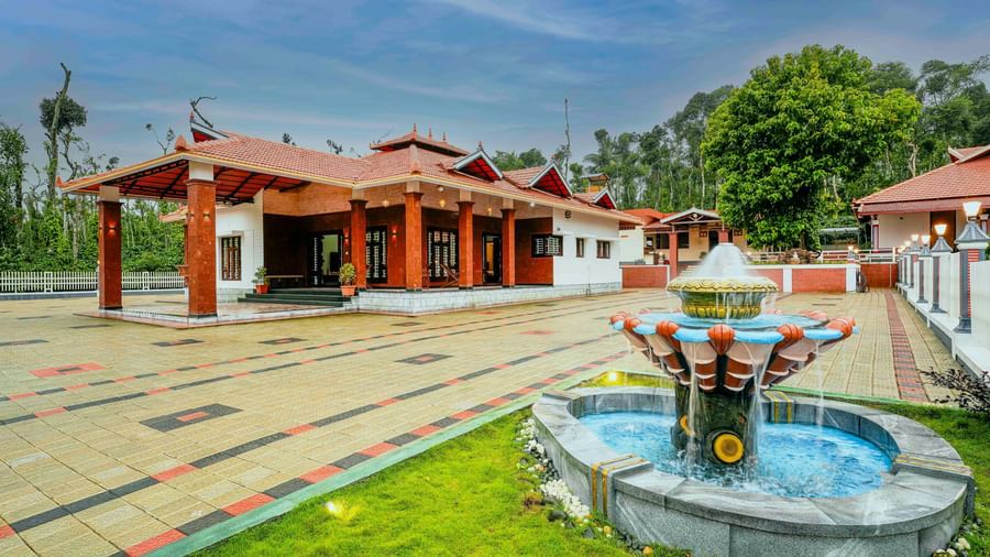 A wide-angle view of the central courtyard at Bhuthanakadu Retreat, featuring a decorative fountain and the traditional architecture of the buildings under a bright, cloudy sky.