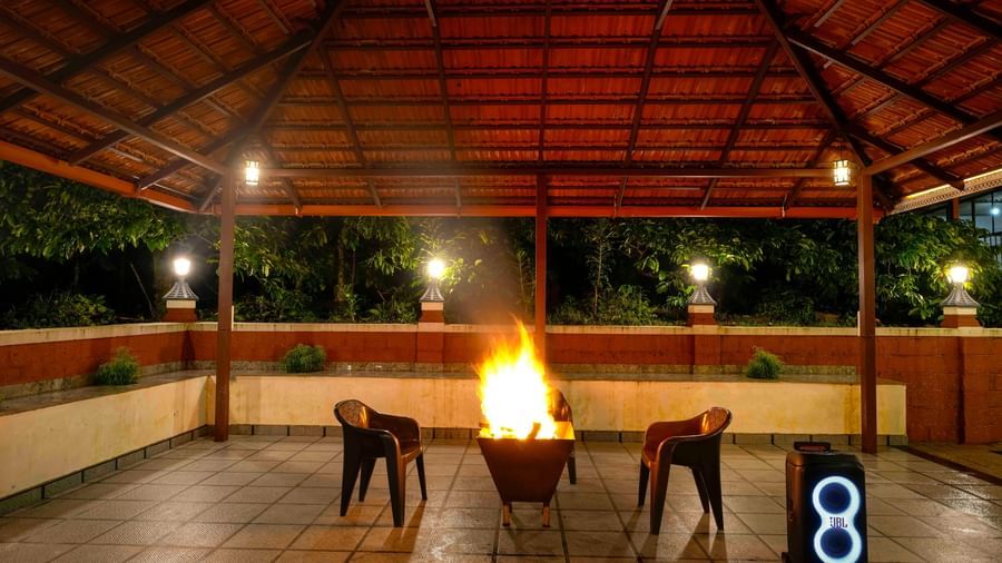 A large bonfire crackles at the center of a covered, open-air pavilion at Bhuthanakadu Retreat, surrounded by chairs under the warm glow of the lights at night.