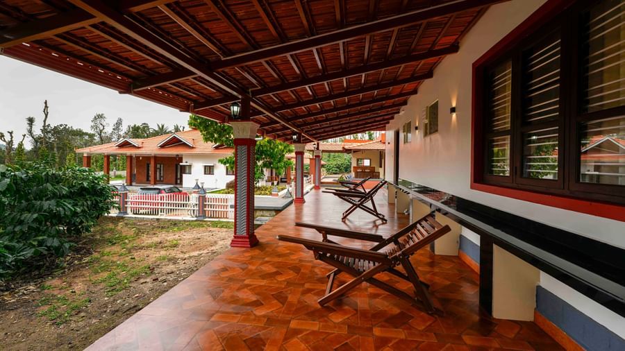 A wide, low-angle shot of a building at Bhuthanakadu Retreat, highlighting a wooden porch with seating and a high-pitched thatched roof.