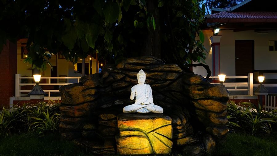 A serene Buddha statue sits on a rock illuminated by a soft, yellow light in a lush garden at Bhuthanakadu Retreat, with buildings visible in the background.