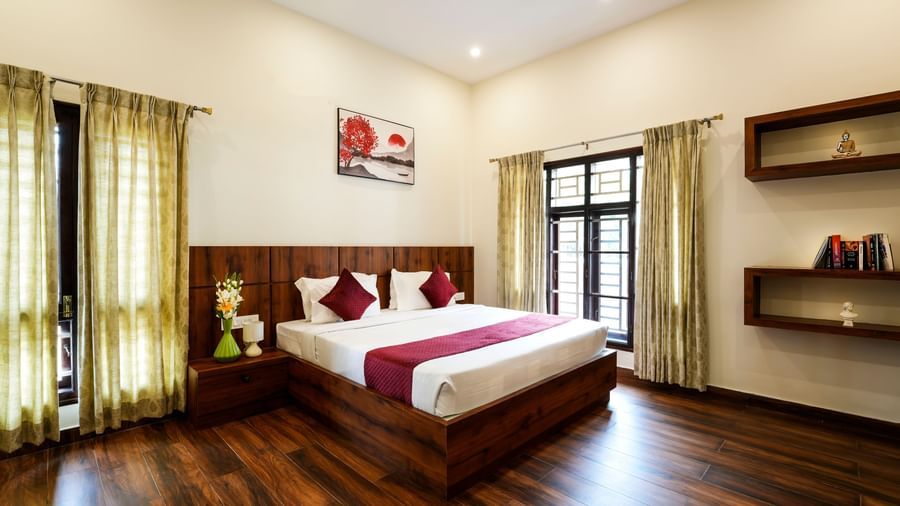  A spacious deluxe room at Bhuthanakadu Retreat, featuring a large bed with a rich wooden headboard, a red and white duvet, and warm recessed lighting.