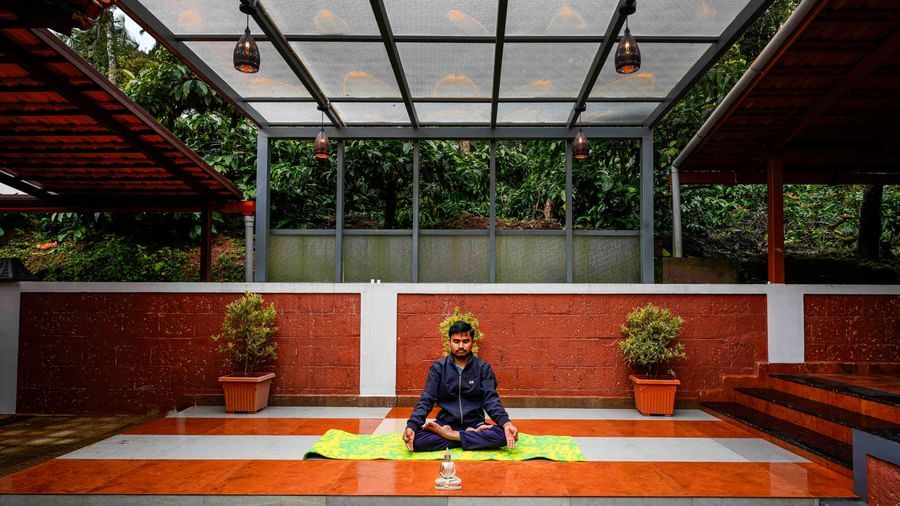 An individual is meditating in a serene, partially covered outdoor yoga space at Bhuthanakadu Retreat, surrounded by lush greenery and red-tiled walls.