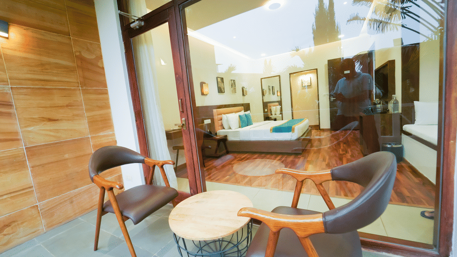 An outdoor patio balcony at Bindiga Peak Resort with 2 chairs and a small table sitting next to a large glass wall that looks into a bedroom.