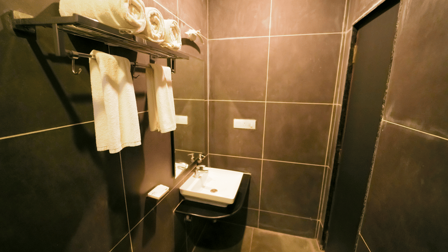 A bathroom with dark grey wall tiles, a white square washbasin, a mirror, and a metal towel rack with white towels at Bindiga Peak Resort.