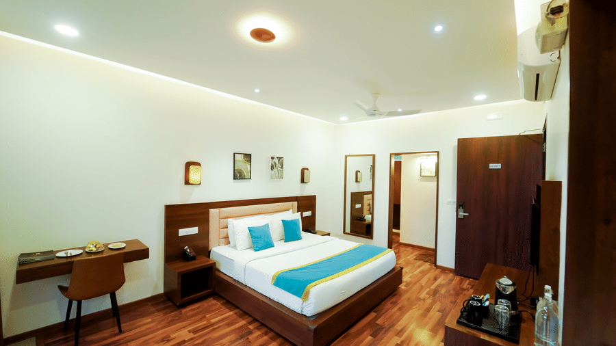 A bedroom with white walls, a large bed, a small wooden workspace, and an open doorway leading to a hallway at Bindiga Peak Resort.