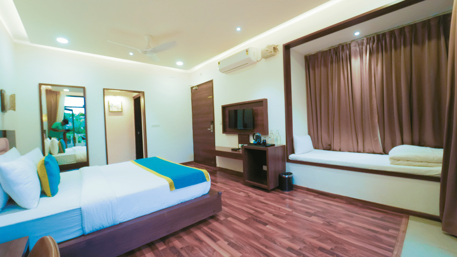 A bedroom at Bindiga Peak Resort with wood-patterned flooring, a wall-mounted TV, and a cushioned bench below a brown-curtained window.