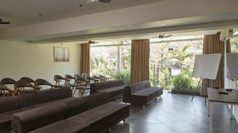 An indoor conference room with rows of brown sofas, chairs, a whiteboard, and glass windows showing outdoor greenery at Bindiga Peak Resort.