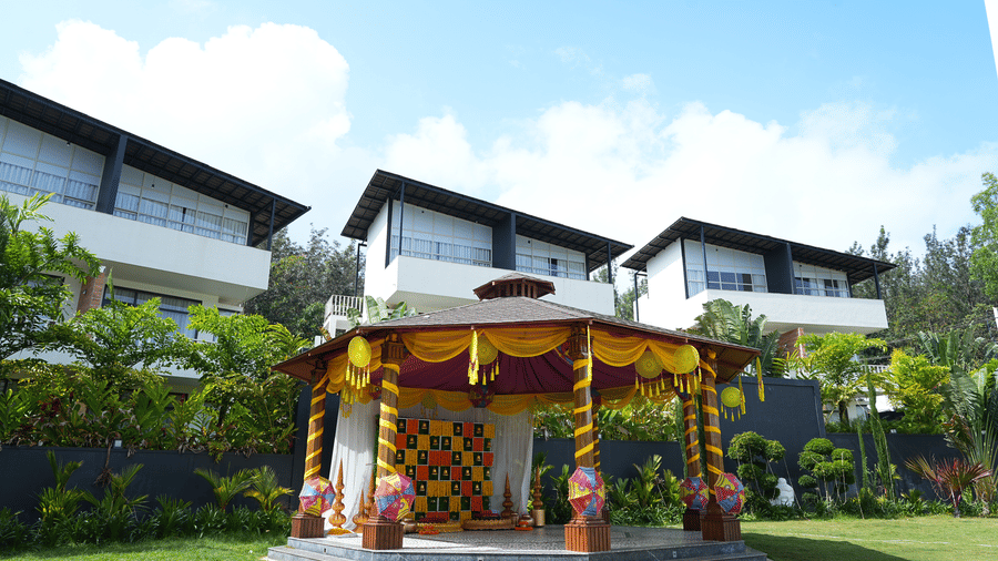 A wooden gazebo decorated with orange and yellow flower garlands on a lawn with white villas behind under a blue sky at Bindiga Peak Resort.