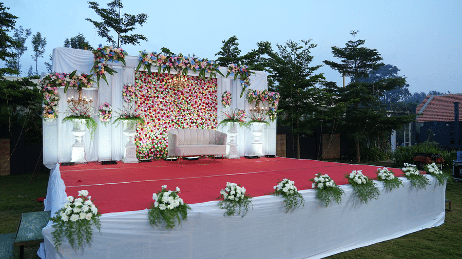 A raised outdoor event stage with a red carpet, white fabric skirting, and floral decorations under a dusk sky at Bindiga Peak Resort.