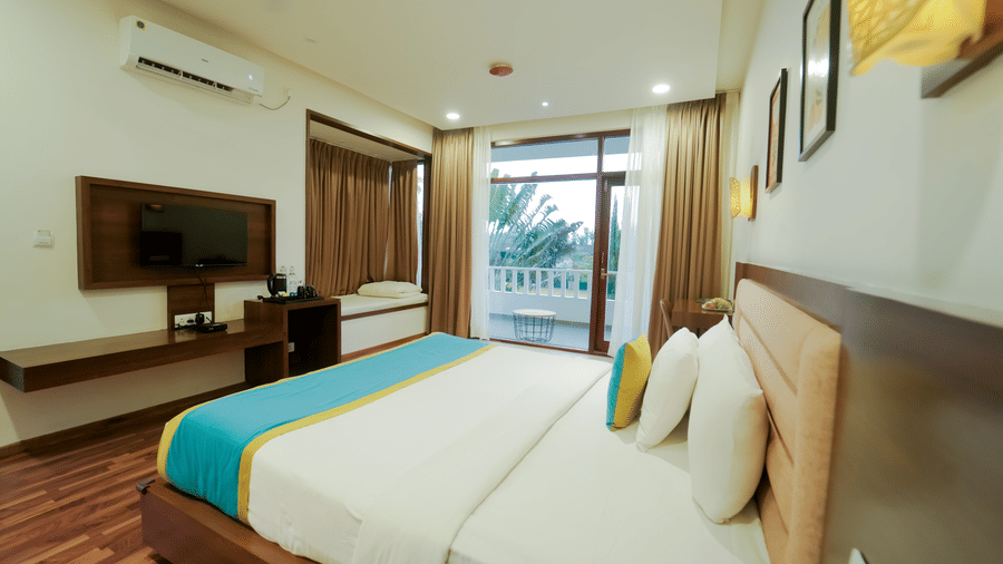An air-conditioned room at Bindiga Peak Resort with a large bed, wooden flooring, a wall-mounted TV, and glass doors leading to the balcony.