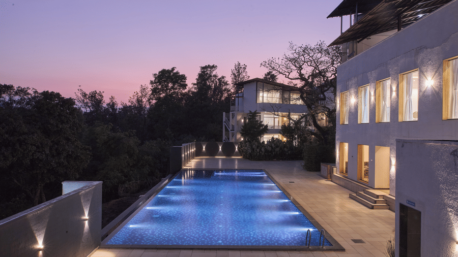 A long outdoor swimming pool flanked by multi-storey white buildings and trees under a pink and purple dusk sky at Bindiga Peak Resort.