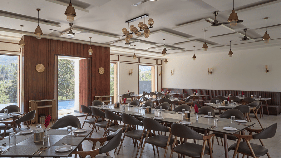 A restaurant featuring wooden tables and chairs, white walls, ceiling fans, and large glass windows at Bindiga Peak Resort.