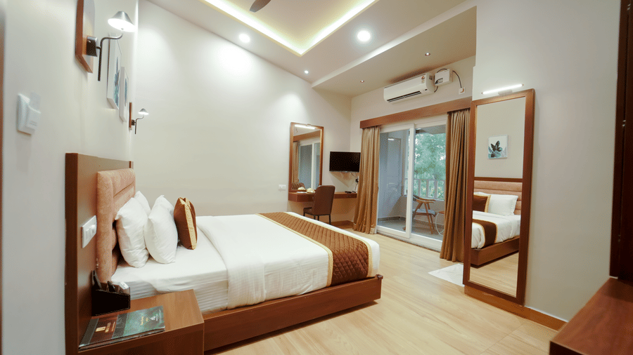 A bedroom with a large bed, light wood-patterned flooring, wall mirrors, and a glass door that opens to the balcony at Bindiga Peak Resort.