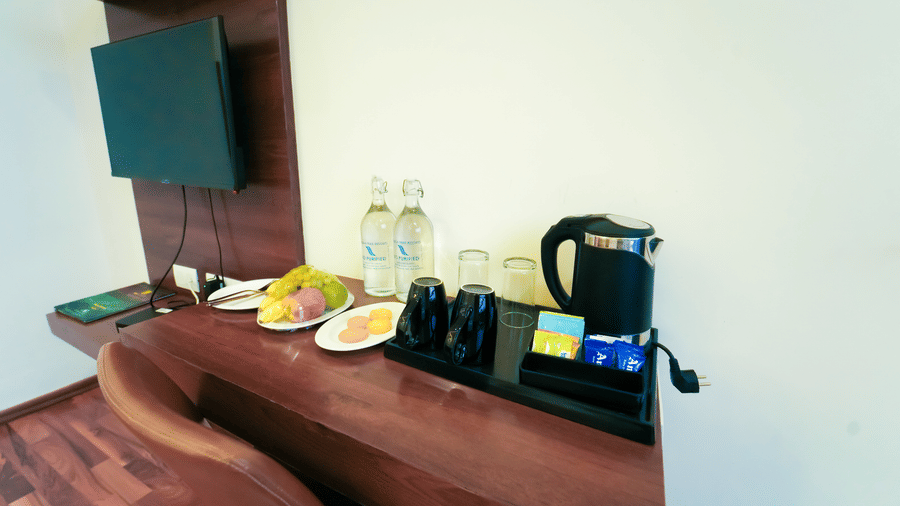A wooden desk with a brown chair, water bottles, an electric kettle, and a wall-mounted television above at Bindiga Peak Resort.
