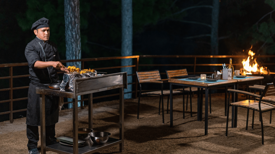 A chef stands at a cooking station on an outdoor terrace at BluSalzz Escapade Resort, Lansdowne, at night. A dining table with chairs and a fire are visible nearby.