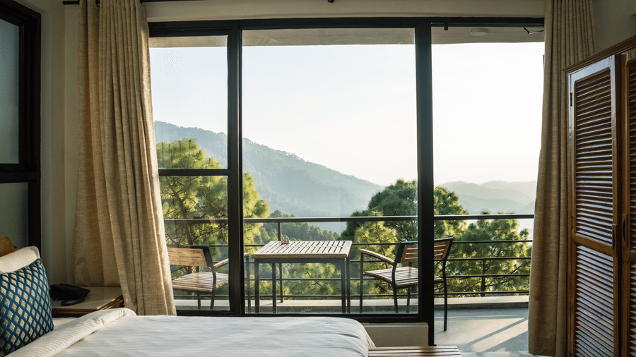 A bed is positioned next to large glass doors that open onto a balcony in the Pine Forest Room at BluSalzz Escapade Resort, Lansdowne. A table and chairs sit on the balcony overlooking hills and trees.