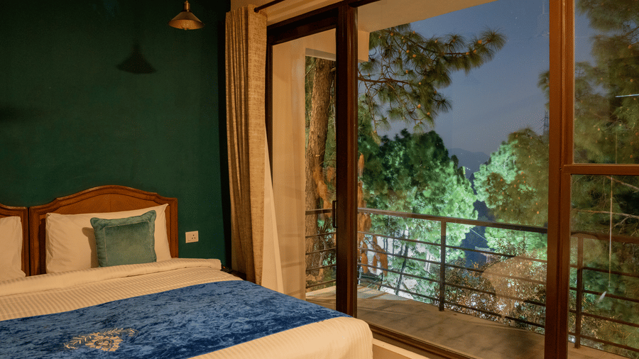 A bed with pillows and a runner sits in the Pine Forest Suite. Large glass doors open onto a balcony at night, showing lit trees outside.