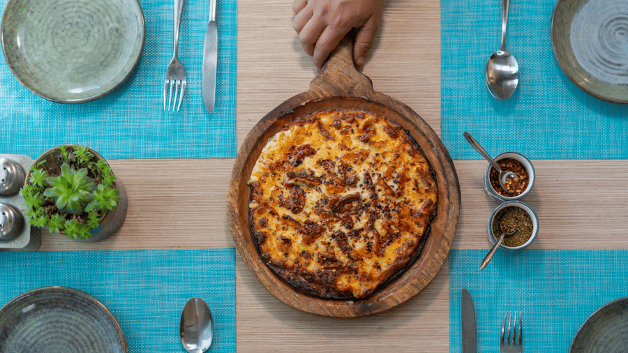 A pizza sits on a board on a dining table set with plates, cutlery, bowls, and a plant at BluSalzz Escapade Resort, Lansdowne.
