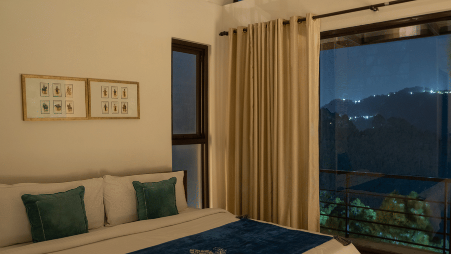 The Sun Terrace Room at BluSalzz Escapade Resort, Lansdowne, features a bed positioned next to a large window at night. Lights from buildings on distant hills are visible outside.