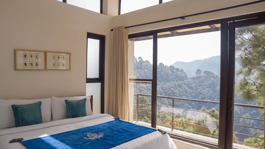 A bed faces a large glass window and balcony door in the Sun Terrace Room at BluSalzz Escapade Resort, Lansdowne. The window overlooks hills and trees under a clear sky.