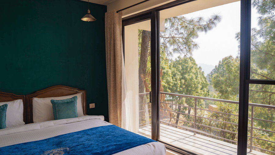 A bed is positioned against a wall next to glass doors that open to a balcony in the Sun Terrace Room at BluSalzz Escapade Resort, Lansdowne. Trees are visible through the glass.