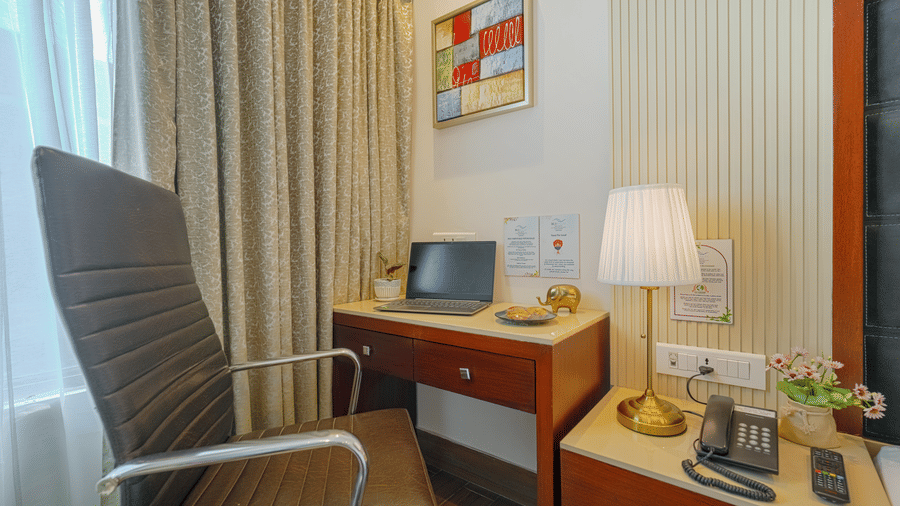 A close-up of a wooden hotel desk with a laptop, lamp, stationary, and a brown leather chair at BluSalzz Select City Centre, Amritsar.