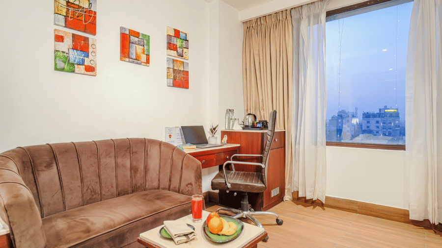 A small sitting and working area in a hotel room with a brown velvet sofa, a laptop on a desk, and colourful wall art at BluSalzz Select City Centre, Amritsar.