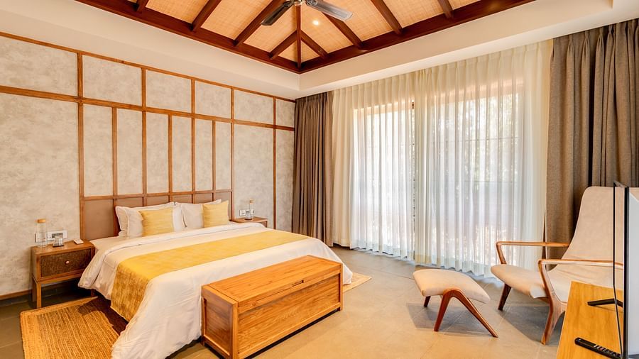 A cosy bedroom with a pitched wooden ceiling, a king-sized bed, warm tones, and soft natural light with  a large window in the background paired with semi transparent curtains  impressions of sunlight - BluSalzz Villas - Alocassia, Anjuna