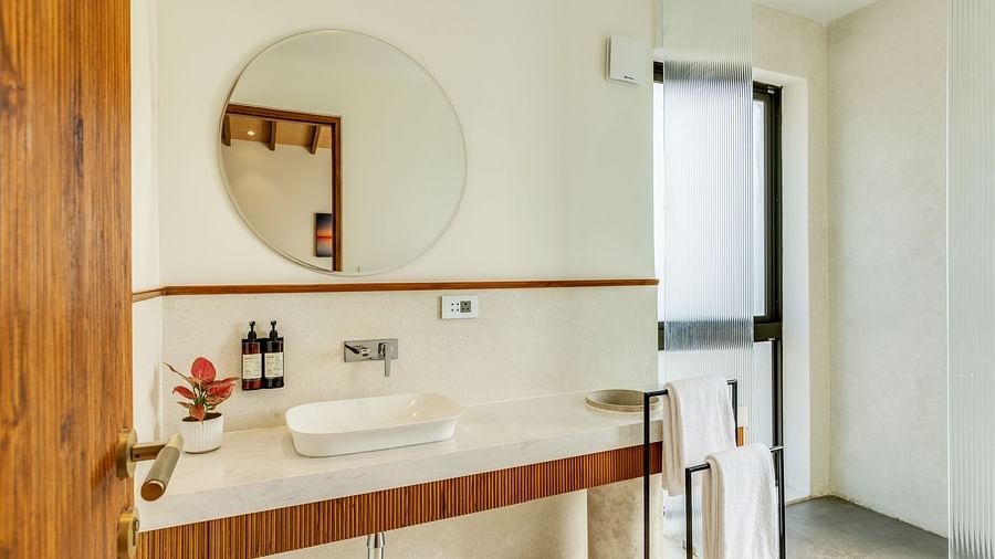 A modern bathroom with a wooden vanity, round mirror, and minimalist fixtures, lit by natural light from a window - BluSalzz Villas - Alocassia, Anjuna