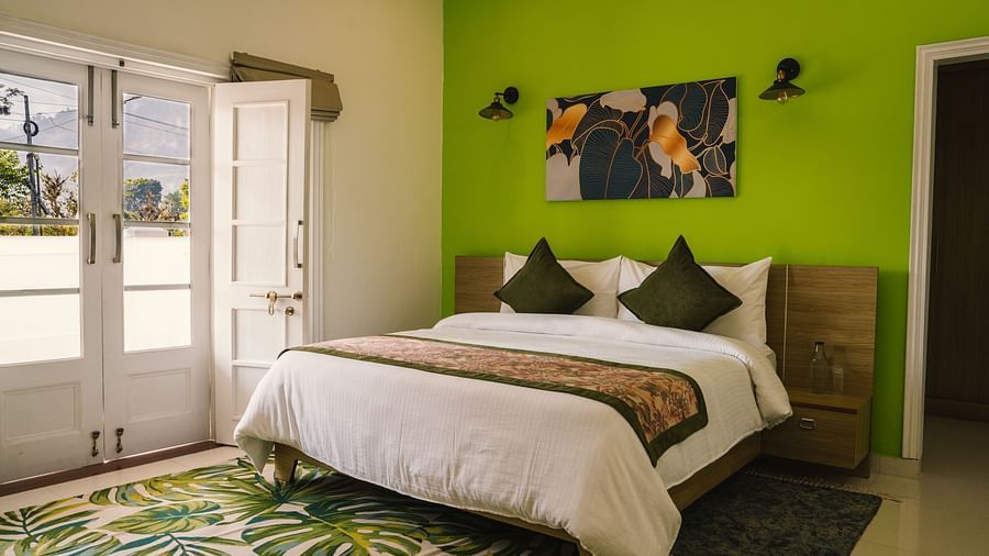A spacious room at BluSalzz Villas - The Green Roof, Bhimtal featuring a large bed, a photo frame above the headboard, a carpet, and a door leading to the balcony.