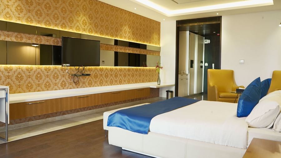 A well-appointed hotel room at BNGV Grandeur Hotel and Banquets, featuring a bed with blue and white linens, a patterned wall, and a dresser with a mirror.