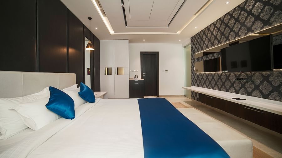 A modern hotel room at BNGV Grandeur Hotel and Banquets, featuring a comfortable bed with a blue bed runner, clean white linens, and sleek furnishings.