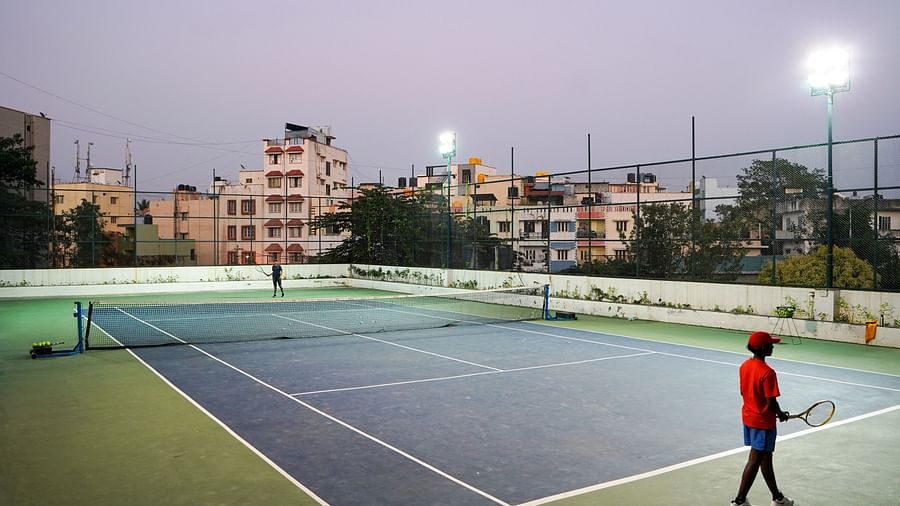 An outdoor tennis court under evening lights with a single player on the court, at BNGV Grandeur Hotel and Banquets.