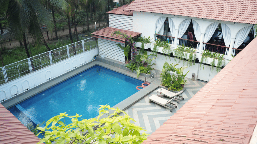 A pool shows an outdoor chequered deck, surrounding trees from a tiled roof perspective at Bougainvillea, Alibaug.